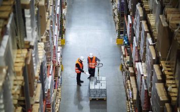 Employer in helmet and uniform showing at packed goods on forklift truck being pushed by worker